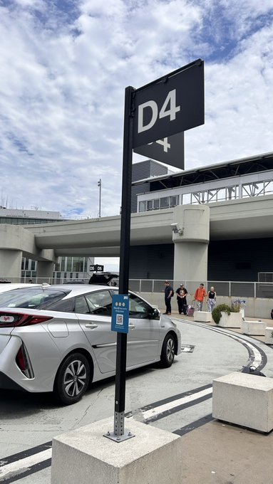 A white Toyota Prius is parked at an airport pickup area under a black sign marked 'D4' with an airplane symbol, while a few people wait with luggage near the terminal.