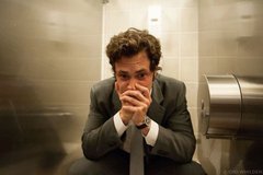 A young man in a gray suit sits hunched over on what appears to be a toilet in a bathroom, head down with hands clasped together near his face in a contemplative or distressed pose. The image visually echoes the emotional weight of heartbreak mentioned in the post, paired with the song "Oxlade Dkt" by the user expressing vulnerability.