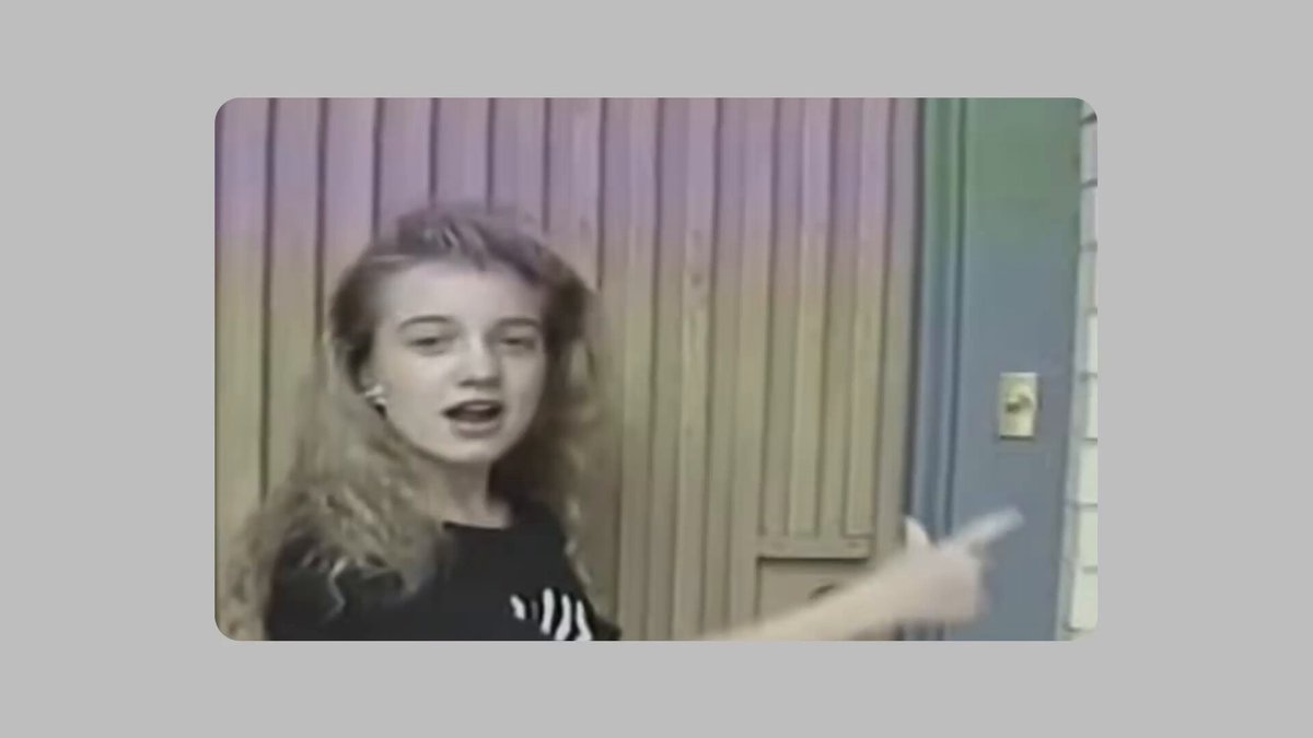 The image features a woman standing next to a closed door, giving a speech. She has long blonde hair that flows down her back. The background of the picture consists of a gray wall with a wooden paneling pattern. There are no texts or other objects visible in this particular image, and it is just the woman's face captured against the backdrop.