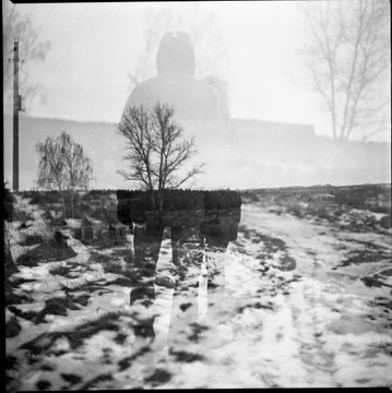 A moody, black and white image showing the ghostly silhouette of a figure in a landscape with trees, with a ghostly stone structure in the foreground.