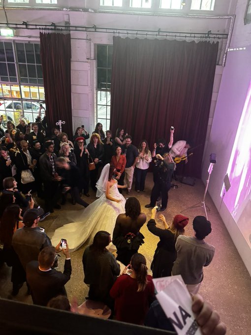 A bride in a flowing white gown and veil stands at the center of an art gallery surrounded by a crowd of attendees, with someone holding up a book or program displaying the letters "NA" while another person appears to be playing a guitar nearby. The scene captures an unconventional wedding ceremony that aligns with the post's message about committing to art in a creative, unconventional way.