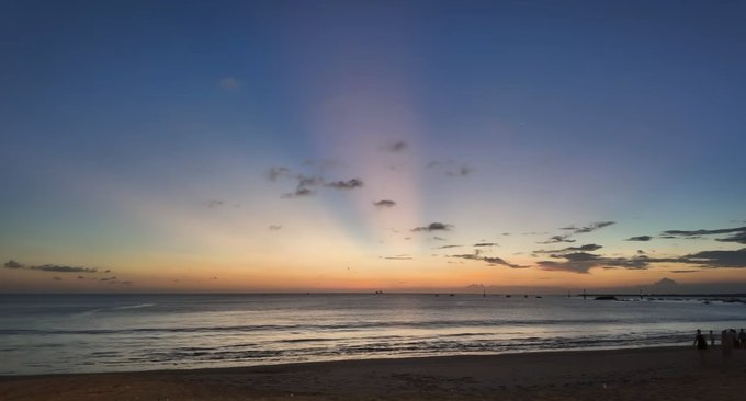 A serene beach scene at twilight, featuring calm ocean waves and a sky illuminated by sunset hues and crepuscular rays.