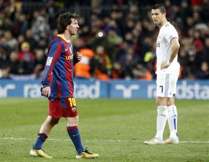 A soccer match scene featuring Lionel Messi in a Barcelona jersey (#10) walking on the field while Cristiano Ronaldo in a white Real Madrid jersey (#7) stands in the background.