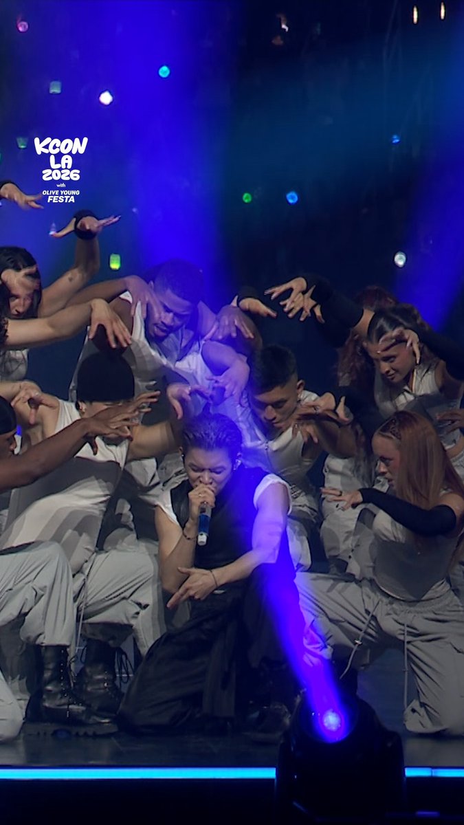 Yeonjun of TXT kneels at the center stage while surrounded by backup dancers in coordinated black-and-white outfits, performing "Coma" at KCON LA 2026, as indicated by the "KCON LA 2026" logo in the upper left corner of the dramatic blue-lit performance.
