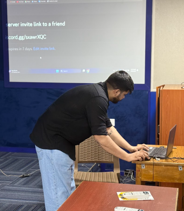 A man in a black shirt and light blue jeans leans over to type on a laptop at a wooden desk, with a projection screen behind him displaying a Discord server invite link 'discord.gg/sxawrXQC'.