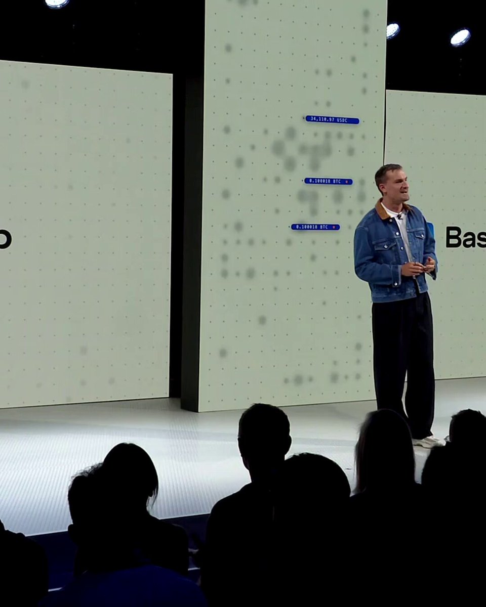 The image shows a man standing at a podium, giving a speech or presentation to an audience of people who are sitting behind him. The speaker appears to be wearing a blue jacket and has his hands up as if he's gesturing during his talk. There is also a screen in front of the speaker that displays some text related to the event.