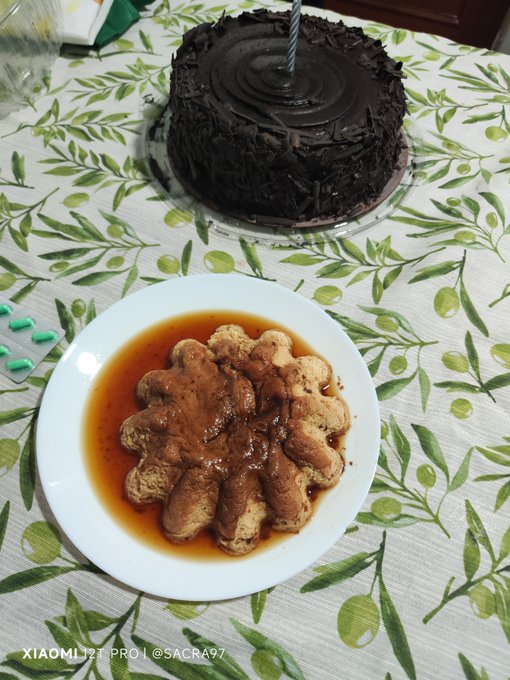A chocolate cake with a striped candle and a plate of star-shaped food drizzled with brown sauce sit on an olive-patterned tablecloth, with a watermark reading 'XIAOMI 12T PRO | @SACRA97'.