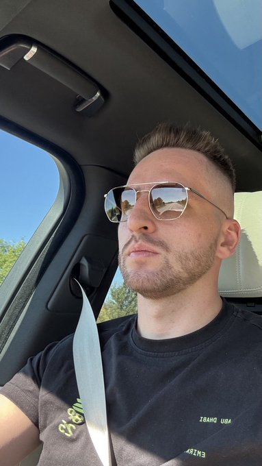 A selfie of a man wearing sunglasses and a black t-shirt sitting in a car with the seatbelt on, reflecting a desert landscape in his glasses.