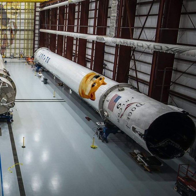 A SpaceX Falcon 9 rocket is displayed inside a hangar with a large Dogecoin dog face logo on its side, along with the 'DOGE-1' mission label and American flag.