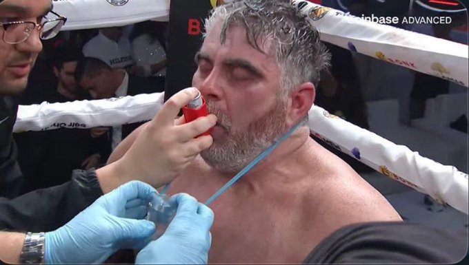 A sweaty boxer receives medical attention in the corner of a boxing ring, with a cornerman holding a red inhaler to his mouth while wearing blue gloves. The 'coinbase ADVANCED' logo is visible on the ring.