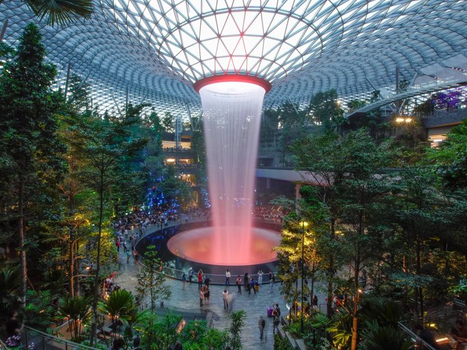 The Rain Vortex, a 40-meter indoor waterfall at Singapore's Jewel Changi Airport, illuminated in pink beneath a glass dome surrounded by lush greenery and visitors. The image showcases Singapore's iconic landmark, tying into the Token2049 conference's theme of exploring cutting-edge crypto innovations and institutional growth in the city.