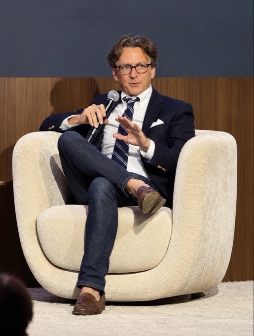 A man in a dark blazer, white shirt, striped tie, and jeans sitting in a cream-colored armchair, speaking into a microphone with his legs crossed.