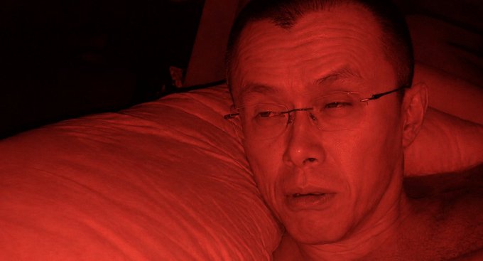 A close-up photo of a man wearing glasses lying on a white pillow, with the entire scene bathed in intense red lighting.
