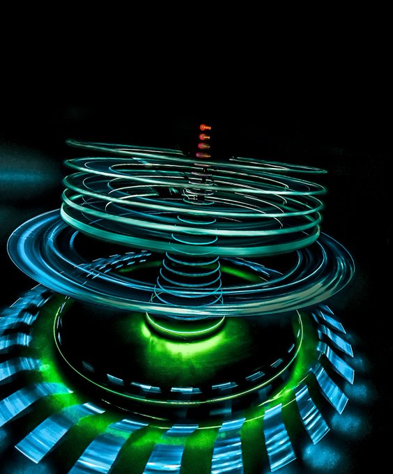 The image is a close-up of a large, spinning light sculpture with blue, green, and red colors, set against a black background at night. The colors are vibrant and illuminated by white lights, creating an atmospheric and visually striking piece. The photo appears to be taken using the light painting technique as there's also text in the image that seems to be related to "Gm! Happy TezosTuesday!".