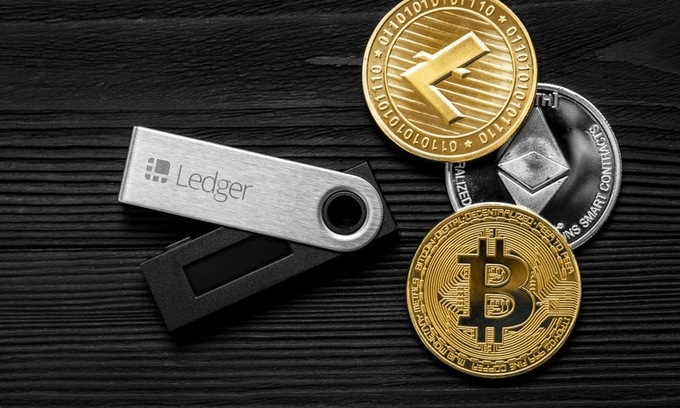 A Ledger hardware wallet sits on a dark wooden surface beside physical representations of cryptocurrency coins, including Bitcoin, Ethereum, and what appears to be a smart contract token. The image visually ties to the post's story about a Ledger user losing nearly $2 million due to a 2020 email list leak, illustrating the hardware wallet brand at the center of the $500 million class-action lawsuit.