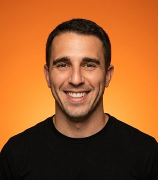 The image features an adult male smiling for the camera against an orange background. The man's face is the main focus of the photo, with his smile radiating warmth and happiness. The surrounding environment appears to be a simple orange color that contrasts nicely with the man's black t-shirt. There are no other objects or people in the image, making it a clear representation of Anthony Pompliano, who has been described as "the percentage of Bitcoin held at a loss is now bigger than those in profit". The post seems to be related to cryptocurrency and the market trends surrounding it.
