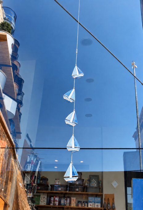 A string of decorative crystal/glass sailboat ornaments hanging in front of a storefront window with a blue reflection, with a wooden shelving unit visible inside.