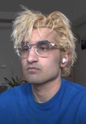 A young man with curly blonde hair, clear glasses, and a blue shirt looks directly at the camera.