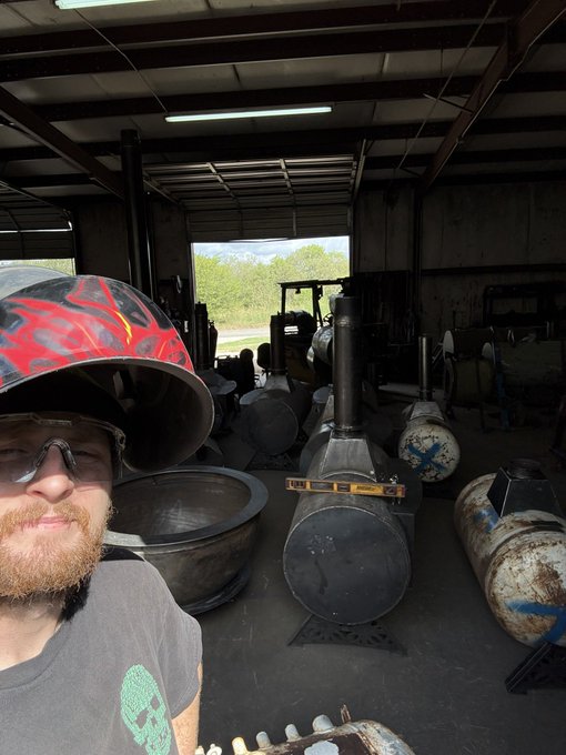 A selfie of a man wearing a red and black flame-patterned welding helmet, glasses, and a black skull t-shirt inside a workshop filled with industrial metal tanks and equipment.