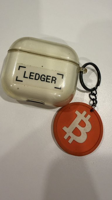 A transparent LEDGER hardware wallet case sits on a white surface, attached to an orange keychain featuring the Bitcoin "₿" symbol, symbolizing a self-custody Bitcoin storage solution amid the criticism Ledger once received from crypto purists for supporting multiple cryptocurrencies.