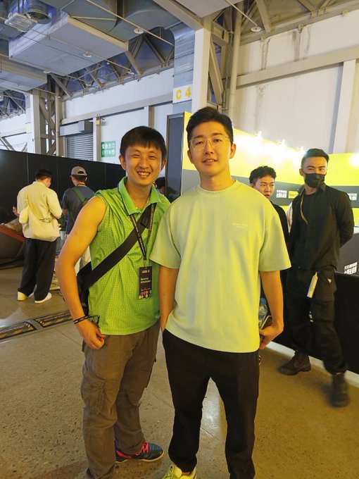 A photograph of two smiling young Asian men posing together at what appears to be a convention or exhibition venue, with one wearing a bright green sleeveless shirt and a lanyard, and the other in a light green t-shirt and glasses.