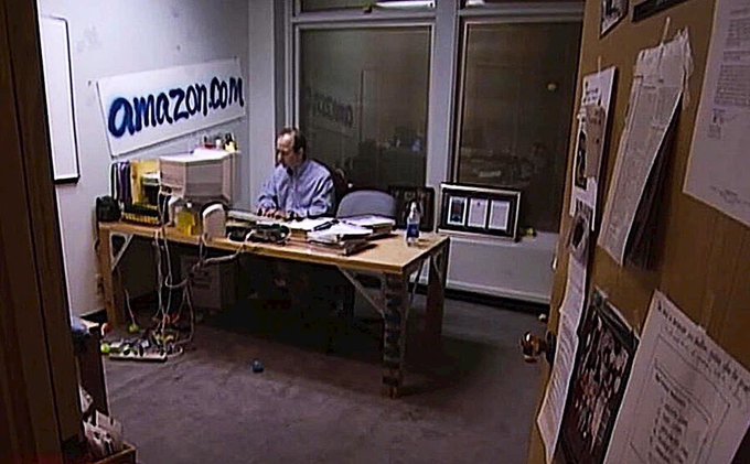 A photo of Jeff Bezos sitting at a desk with an early 'amazon.com' banner on the wall behind him, surrounded by computer equipment and cardboard boxes.