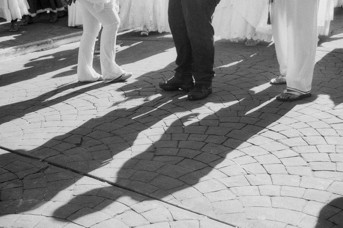 A black and white photograph showing the lower legs and feet of several people, casting long dramatic shadows across a cobblestone courtyard, with white fabric visible in the background.