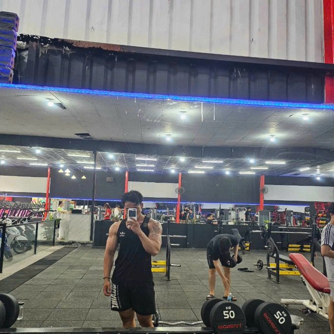The image shows a man taking a selfie with his dumbbells, likely at a gym or fitness center. The man is standing next to a weight rack while holding a pair of dumbbells in front of him. There are other people present in the scene as well, indicating that this might be a popular location for working out and socializing.