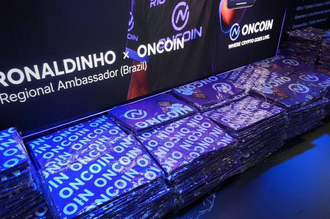 Stacks of blue "ONCOIN" branded merchandise sit on a table beneath a backdrop featuring "RONALDINHO x ONCOIN Regional Ambassador (Brazil)" signage and the tagline "WHERE CRYPTO GOES LIVE," tying directly to the post's praise for OnCoin's standout TOKEN2049 booth and its free merch giveaways.