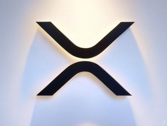The XRP (Ripple) cryptocurrency logo—a stylized black X—mounted on a white wall.