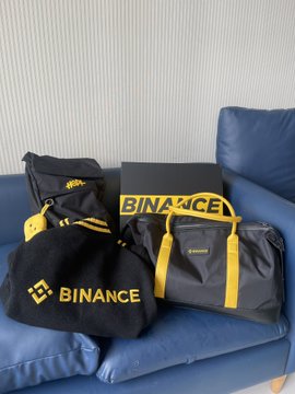 Black and yellow Binance-branded merchandise including bags and pouches are displayed on a blue couch.