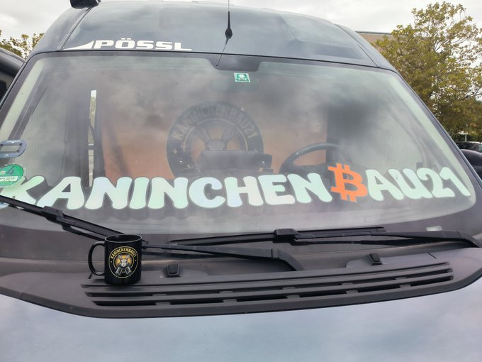 The front windshield of a Pössl van with 'KANINCHENBAU21' decal incorporating a Bitcoin symbol, plus a black mug with the same logo resting on the hood.