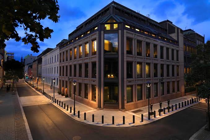 A modern multi-story office building with illuminated windows stands at a street corner during twilight, surrounded by pedestrian walkways and bollards. The image appears unrelated to the post's content about Norway's sovereign wealth fund (NBIM) revealing its first investment in BitMine Immersion Technologies, an Ethereum treasury company, as the building shown could represent the fund's headquarters but lacks any visible identifying text or logos to confirm this connection.