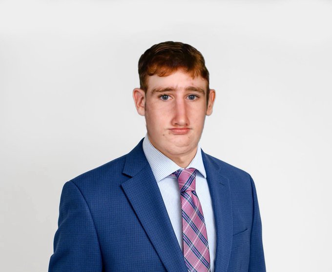 A portrait photo of a young man with reddish hair, wearing a blue suit jacket, light blue dress shirt, and a pink and blue plaid tie, against a white background.