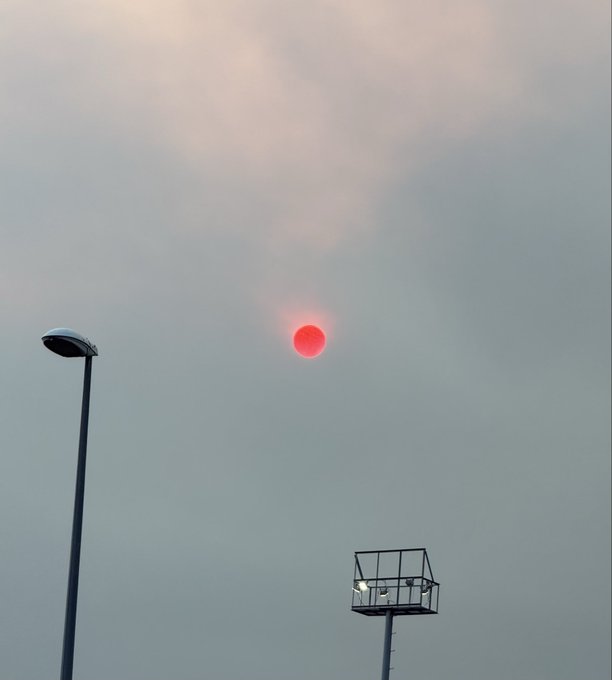 A red sun (likely from wildfire smoke) visible through a hazy sky, with a streetlamp and what appears to be a stadium light fixture in the foreground.