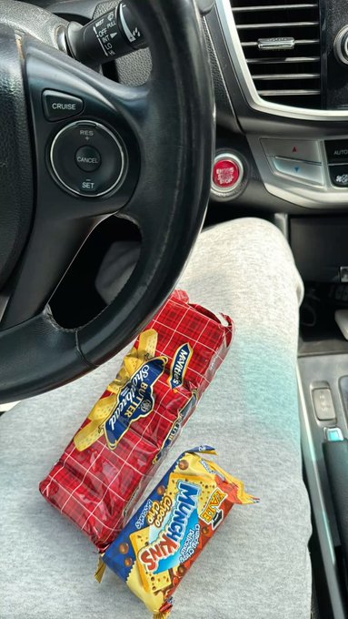 A view from inside a car showing a steering wheel and dashboard, with two packs of MunchyMax biscuits resting on the driver's leg.