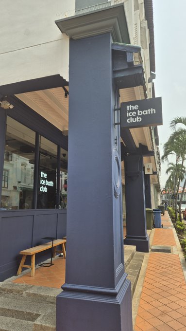 An exterior photo of 'The Ice Bath Club' featuring a dark blue/grey storefront with a prominent pillar and signage, with a wooden bench outside.