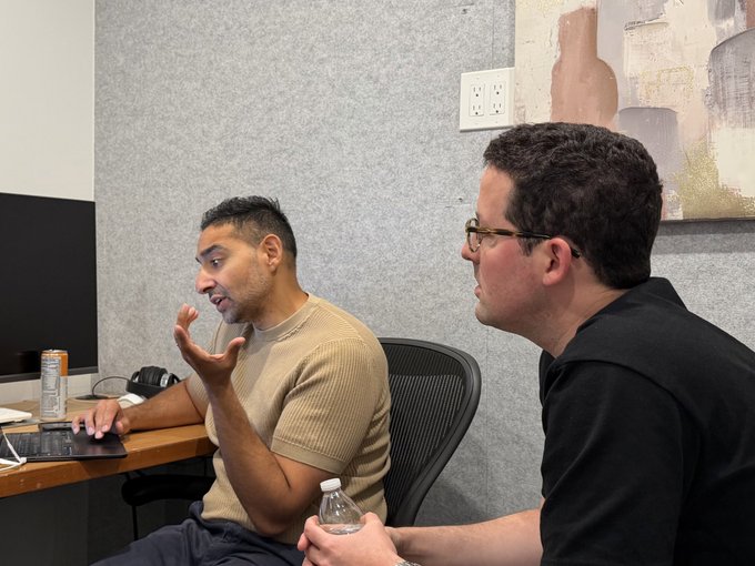 Two men in an office setting; one in a tan shirt gestures with his hand while sitting at a desk with a keyboard, and the other in a black polo holds a water bottle.
