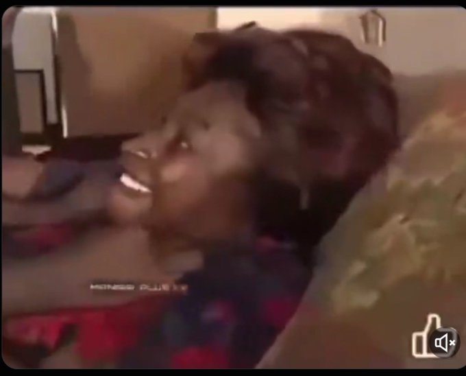 A young person is lying back laughing heartily on what appears to be a couch with floral-patterned cushions, with partially legible subtitles at the bottom of the frame and a muted speaker icon in the corner. The caption "Bitget hack keh... And na just today youkay boi beg for money o" combined with the unrestrained laughter suggests the image captures a moment of uproarious amusement at news of a Bitget crypto hack and a related figure asking for funds.
