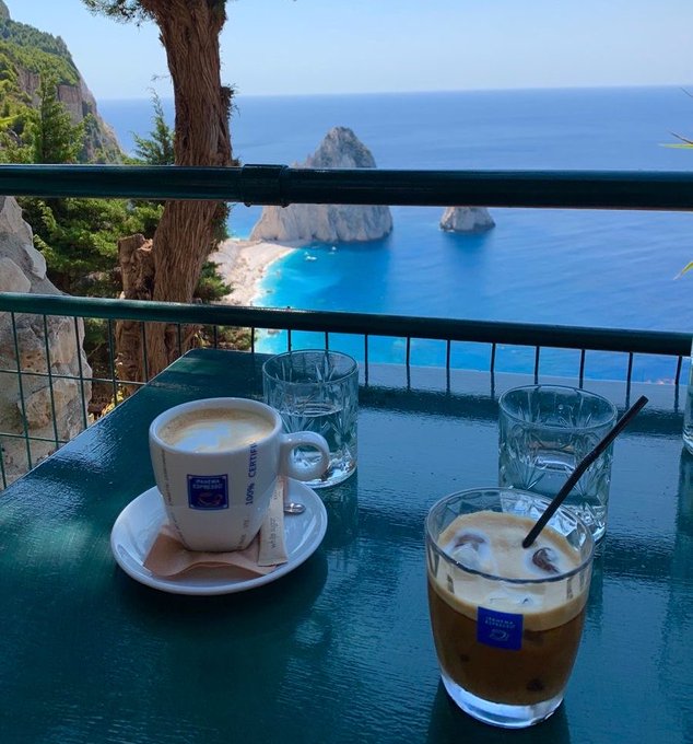 A scenic balcony view overlooking a turquoise sea with rocky formations, featuring a table set with a cappuccino, an iced coffee, and water glasses.