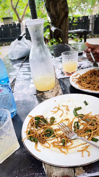 A partially eaten meal of stir-fried noodles with greens on a white plate sits on a wooden outdoor table next to a glass carafe of yellowish drink and a plastic water bottle.