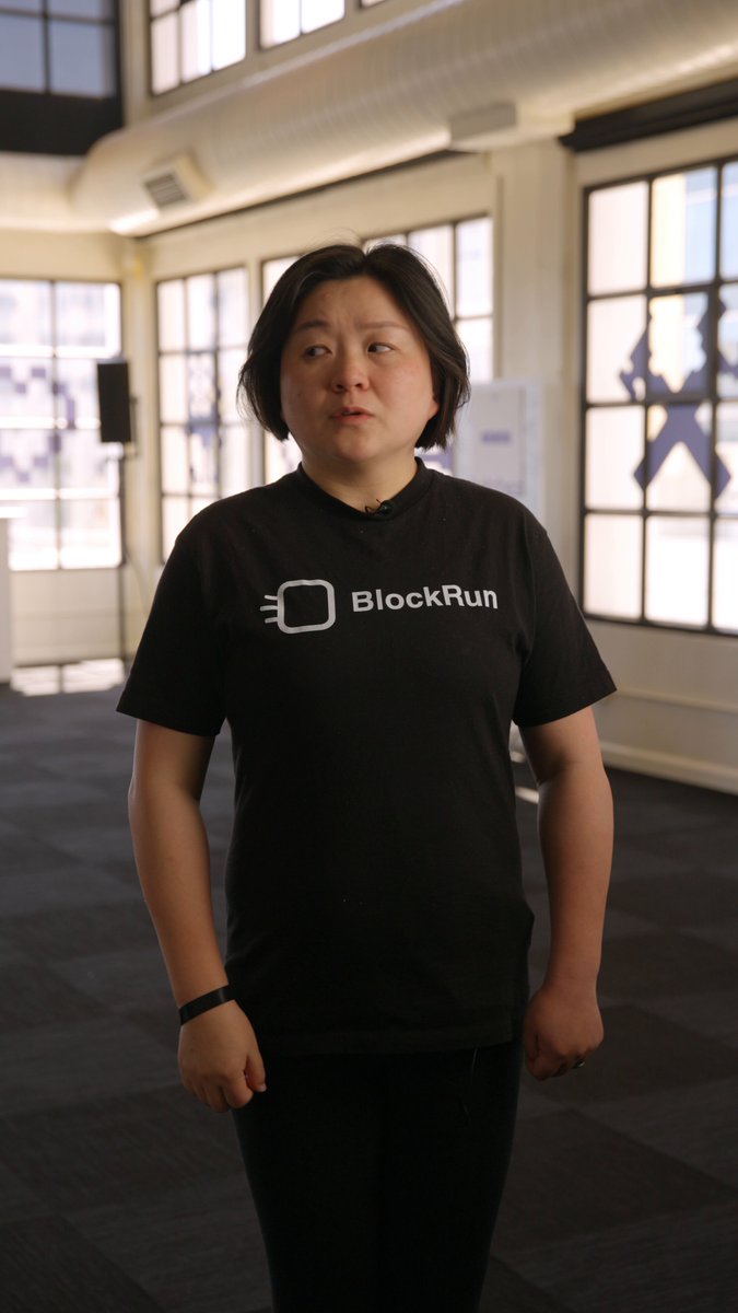 The image features a woman wearing a black t-shirt with white text that says "blockrun" on it, standing in an office setting. The background of the photo shows a window and a speaker system, suggesting that they are in a room where work or meetings take place. There is also a sign visible in the scene, which reads "USDC will be the default payment settlement for agents".