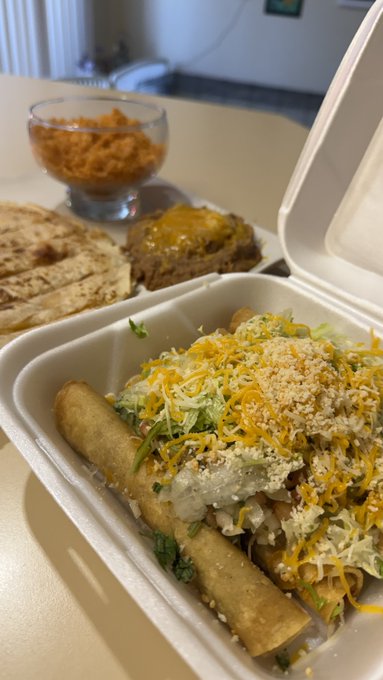 A takeout container holds flautas or rolled tacos topped with shredded lettuce, cheese, crema, and crumbled cheese, with a side of flatbread and a small glass bowl of orange sauce on the table.