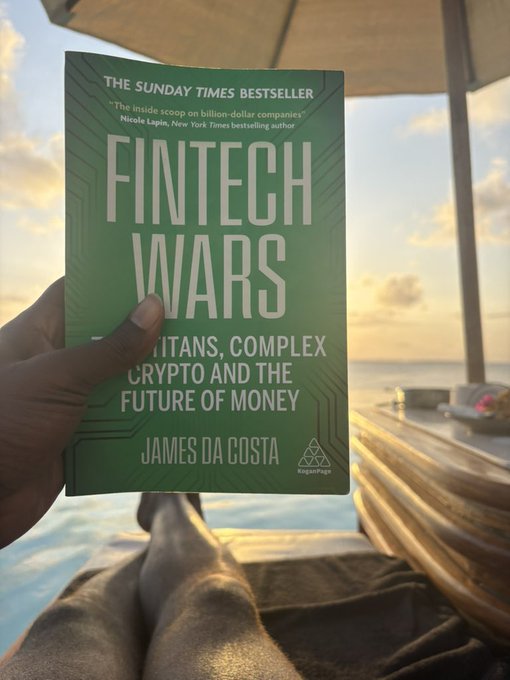 A hand holds the Sunday Times Bestseller book 'FINTECH WARS: Titans, Crypto and the Future of Money' by James Da Costa, displayed against a serene ocean sunset backdrop.