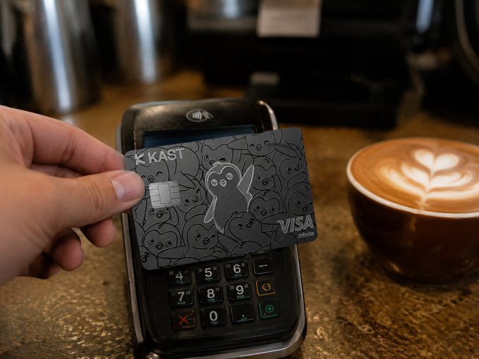 The image shows a person holding a credit card over a black terminal that has a Visa logo on it. The person's hand is reaching for the card, indicating they are about to make a payment using their KAST Card. The scene takes place in front of a coffee cup and a cash register, suggesting that this could be a coffee shop or cafe setting where customers can pay with their credit cards.