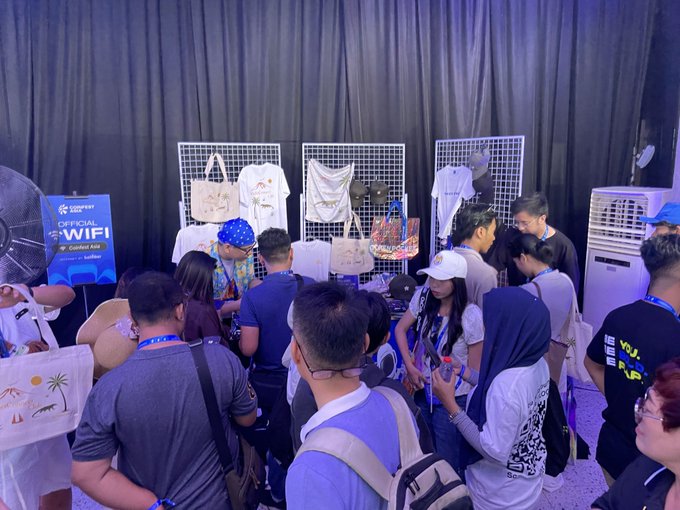 A crowded indoor event where attendees gather at a merchandise booth displaying t-shirts, tote bags, and caps on grid panels, with an 'OFFICIAL WIFI' sign nearby.