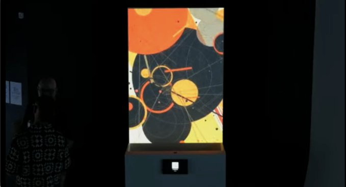 A person stands viewing a Kandinsky-style abstract artwork displayed in a backlit exhibit, featuring black circles and intersecting lines against orange and yellow backgrounds.