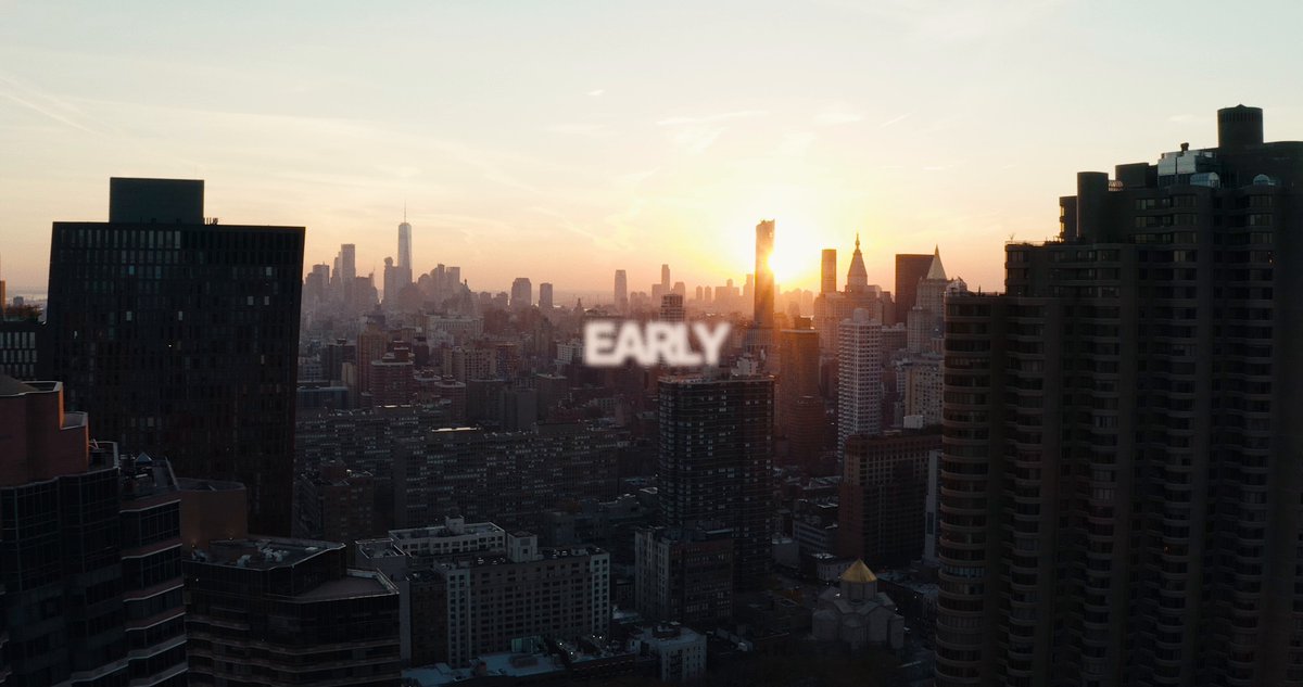 The image shows a city skyline with buildings of various sizes and designs, including skyscrapers and apartment buildings. The sun is setting over the city, casting an orange glow that illuminates the scene. In the foreground, there are two texts written in white letters that read "Early" and "@RWA_xyz". These texts appear to be related to a post or social media account about cryptocurrency.