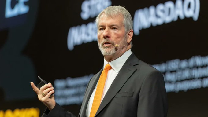The image features an older man wearing a suit with an orange tie speaking into a microphone while sitting at a desk during what appears to be a presentation or speech about AI investing. He has a noticeable gray beard, which adds character to his appearance. The setting suggests that he is addressing an audience in front of a large screen displaying the name "Michael Saylor: 'AI infrastructure is absorbing capital'. This isn't about a single price move". It seems that Michael Saylor is discussing how companies investing in AI are capitalizing on market growth opportunities in this field, emphasizing the increasing importance and potential for returns on investment.