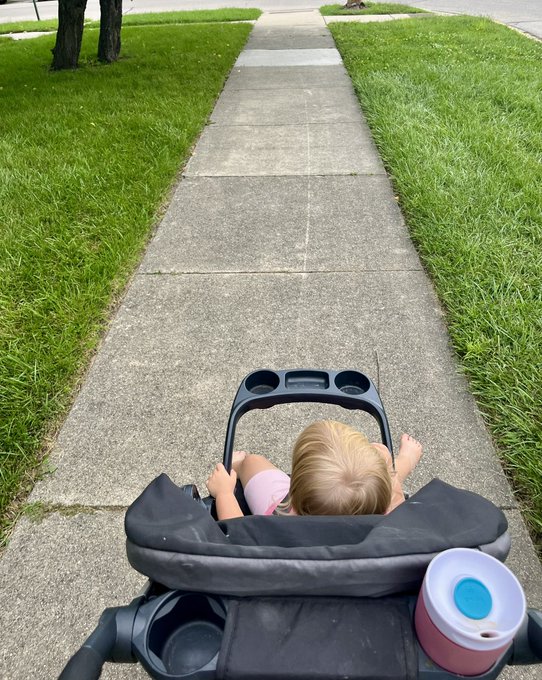 A young child with blonde hair relaxes in a stroller on a suburban sidewalk, with a pink and blue sippy cup beside them, capturing a peaceful morning moment that complements the grateful "Absolutely blessed, Good morning" sentiment.